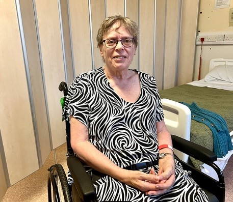 a blond woman wearing a blue and white dress sitting in a wheel chari by a hospital bed