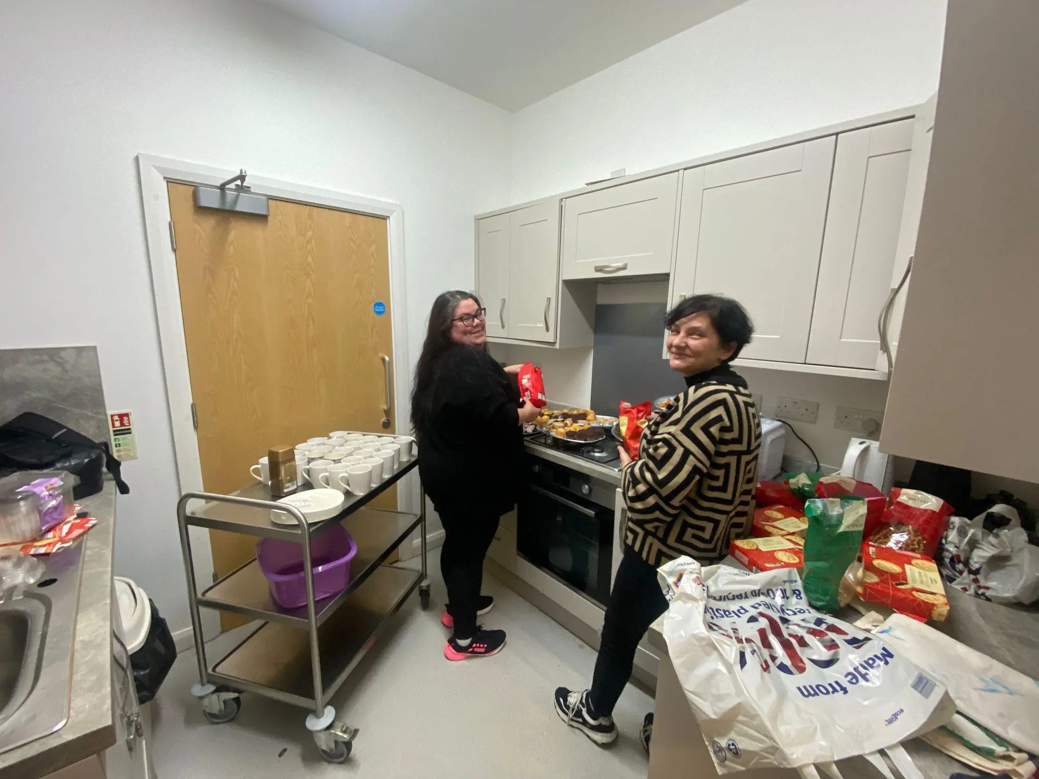 two smiling women in a kitchen sorting food from bag.