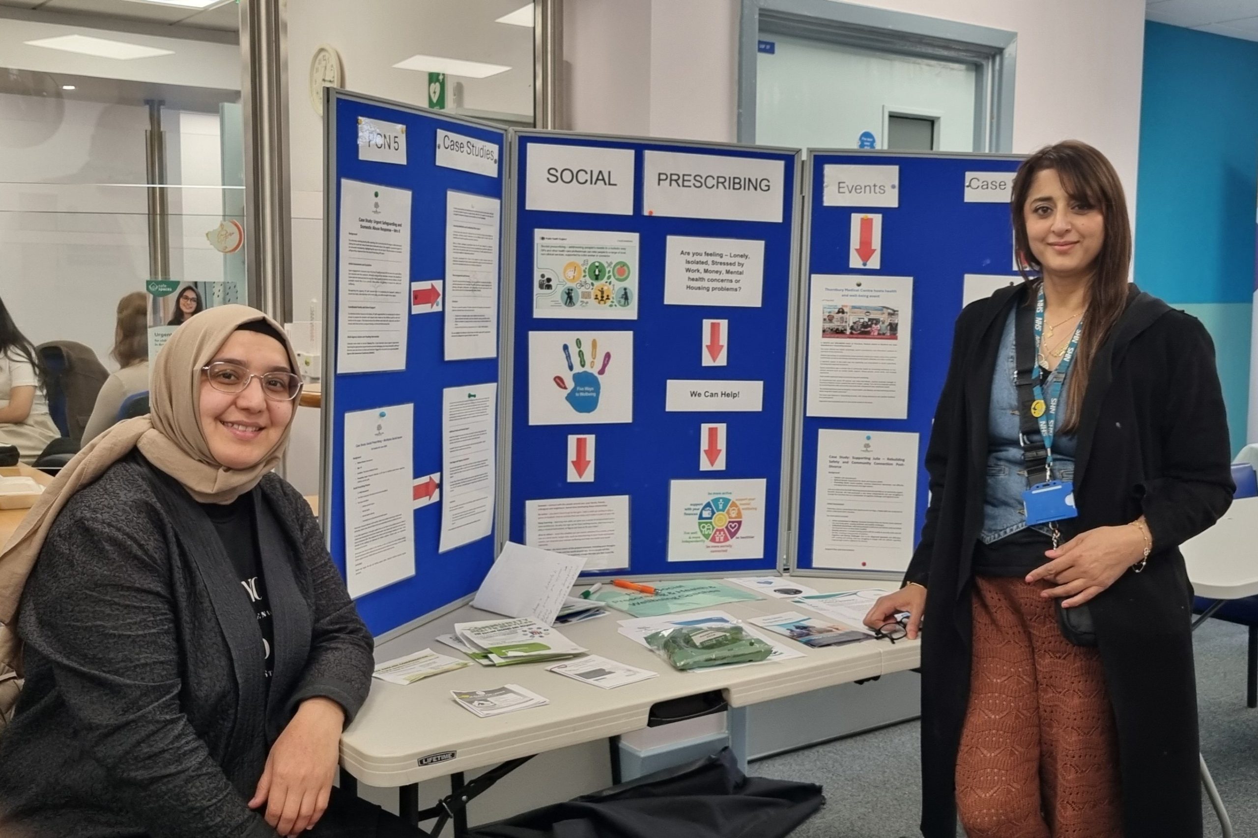 two women with one wearing a hijab sitting by a stand with many posters on a table in Hillside Bridge Medical Centre. They are social prescribers.